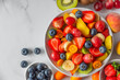 Summer fresh fruit salad in a bowl on white marble background. Top view with copy space. Healthy food for breakfast. Mixed strawberries, grapes, banana, kiwi, blueberries, peach, citrus for diet lunch