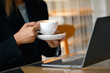 Businesswoman holding a cup of coffee sitting with laptop in modern cafe