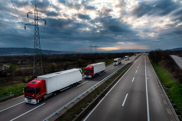 Wall Mural - Convoy of red trucks with white containers on highway, cargo transportation concept in springtime - freight service, with a dramatic sunset sky in background