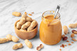 Peanut butter with spoon in glass jar with peanuts in wood bowl on marble table