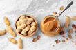 Peanut butter in spoon on jar with peanuts in wooden bowl on marble table