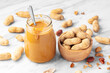 Peanut butter with spoon in glass jar with peanuts in wood bowl on wihite marble table