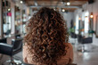 © SOLDATOOFF - Back view of a young Hispanic woman with curly brown hair sitting in a hair salon.