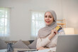 A young Muslim woman wearing a hijab sits contentedly shopping on her laptop, paying through an online banking app and holding a credit card.