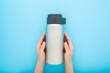 Young woman hands holding and showing closed new silver steel thermos with dark black plastic mug for hot drink or soup on pastel blue table background. Closeup. Point of view shot. Top down view.