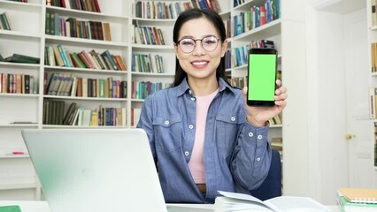 Asian female student showing phone with green screen while sitting at desk in university library. Color key, vertical mockup. Template for advertising. Young woman shows smartphone looking at camera