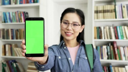 Close up. Asian female student showing phone with green screen standing in campus library space. Color key, vertical mockup. Template for advertising. Young woman shows smartphone looking at camera