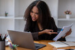 © Wasana - Stressed African American businesswoman reviewing documents while working on a laptop in a modern office.