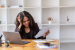© Wasana - Stressed African American businesswoman reviewing documents while working on a laptop in a modern office.