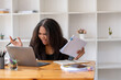 © Wasana - Stressed African American businesswoman reviewing documents while working on a laptop in a modern office.