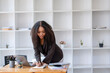 © Wasana - African American businesswoman happily organizing paperwork and using a laptop in a well-lit modern office.