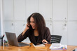 © Wasana - Cheerful African American businesswoman having a phone conversation while working on a laptop in a modern office.