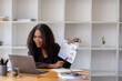© Wasana - Stressed African American businesswoman reviewing documents while working on a laptop in a modern office.
