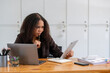 © Wasana - Stressed African American businesswoman reviewing documents while working on a laptop in a modern office.