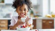 black child having breakfast eating yogurt with fresh fruit, sitting at the kitchen table. Healthy morning routine, breakfast with fresh fruit. Black child enjoying healthy food in the kitchen.