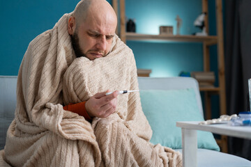 Man wrapping in plaid holding at digital thermometer for measuring body temperature. Sick male with cold and fever