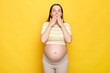 Shocked astonished Caucasian pregnant woman with bare belly wearing casual top isolated over yellow background screaming with amazement covering mouth with hands