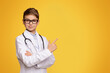 © Prostock-studio - Important information. Serious teenager in doctor uniform pointing aside at empty space, orange studio background