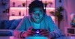 © inthasone - Happy asia man gamer wear headphone competition play video game online with smartphone colorful neon light in living room at night modern house. Esport streaming game online, Home quarantine activity.