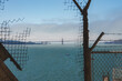 View through rusted bars and wire mesh inside Alcatraz overlooking San Francisco Bay. Golden Gate Bridge visible in distance with boats on calm water.