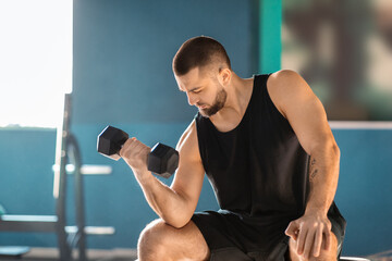 Canvas Print - A man is seated on a bench in gym, gripping a pair of dumbbells in his hands. He appears focused and concentrated on his workout routine, showcasing determination and commitment to fitness.