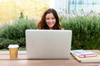 Front view of happy redhead female college student using laptop outdoors sitting on table studying and doing homework.