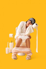 Wall Mural - Sleepy young woman in gown with paper roll sitting on toilet bowl against yellow background