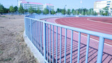 Fototapeta  - Metal fence around the stadium. Closeup of a steel fence surrounding an outdoor sports stadium with a running track on the background of buildings and blue sky with copy space with selective focus.