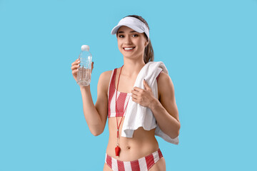 Wall Mural - Young female lifeguard in cap with water bottle and towel on blue background