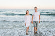 Man and woman walk on water with big waves in sea. Happy couple in love holding hands, looking at camera walking barefoot on seashore. Female and male run on beach ocean. Spending time together