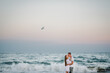 Seagull flies over sea. Couple in love hugging and kissing on seashore. Female kisses and hugs male standing on water with big waves ocean and enjoying summer day. Man embraces woman walking on beach.