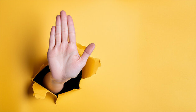 Hand making a stop sign, coming out of a torn yellow paper hole. Concept of stop