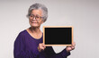 Senior woman holding a mini blackboard and looking at the camera with a smile while standing on a gray background.