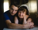 Fototapeta Sawanna - In the portrait, a grandmother smiles warmly as her grandson affectionately embraces her, capturing a timeless moment of love and connection between generations.