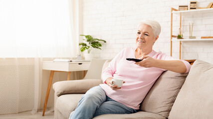 Wall Mural - Elderly woman sitting comfortably on a couch, holding a remote control in her hand. She appears to be focused on the screen in front of her, possibly watching television or streaming content.