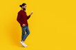 Indian man wearing a red sweater is focused on his cell phone screen, scrolling and reading content. He is standing on yellow studio background, copy space