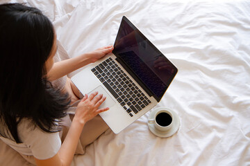 top view of Asian Woman sitting on the bed with cup of coffee and working on her laptop.