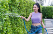 Amidst the lush surroundings of her garden, a woman smiles joyfully as she waters a dense green hedge using a blue garden hose, enjoying the simple pleasures of gardening. Minimal free time leisure.