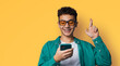 Smiling funny young man wear braces, sunglasses glasses hold typing text look read cell phone cellular smartphone cellphone, crossing fingers, pray hope for good luck isolated orange yellow background