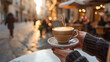 Hands holding a steaming cup of latte in an outdoor cafe setting.