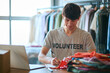 Male Charity Worker At Desk With Laptop Checking Clothing Donations At Thrift Store