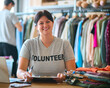 Portrait Of Female Charity Worker At Desk With Laptop Checking Clothing Donations At Thrift Store