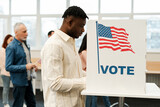 Fototapeta  - Young African American man standing in voting booth with American flag, voting at polling station