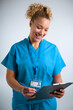 Studio Portrait Of Smiling Female Nurse Wearing Scrubs With Clipboard Against Grey Background