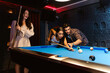 Young diversity group of people playing pool together with smile, enjoyment and fun. Young people spend time in billiards room at the nightclub. Men, women friends playing billiards. Nightlife concept