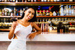 Portrait of an Asian glamour, wearing a white glitter dress smiling and drinking a glass of wine at a luxury nightclub counter bar with liquor in the background. Attractive woman enjoy and fun hangout