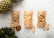 Packages with dried sweet pineapple slices and circles on light background with raw pineapple fruit.Top view.