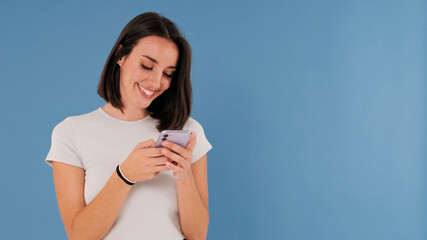 Wall Mural - Beautiful smiling brunette young woman dressed in white top hold using mobile phone isolated on blue background in studio