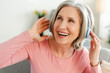 Overjoyed senior woman listening music in wireless headphones, having fun sitting on sofa