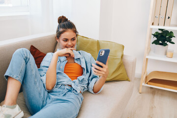 Wall Mural - Happy woman sitting on a cozy sofa, holding a smartphone and smiling while enjoying online gaming at home